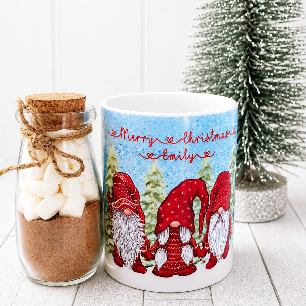 Mug with Christmas design featuring gnomes and a jar of hot chocolate mix on a white surface.