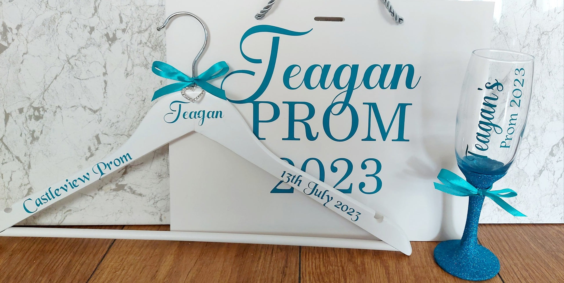 A set of personalized prom gifts including a custom name hanger, a personalized flute glass with 'Prom' and a date, and a gift bag with ribbon decorations.