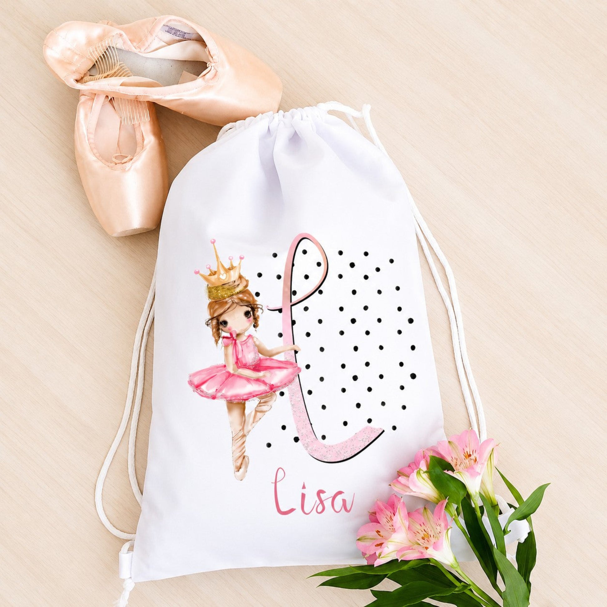 White drawstring bag with a ballerina design and the name 'Lisa', next to pink ballet shoes and flowers on a beige background.