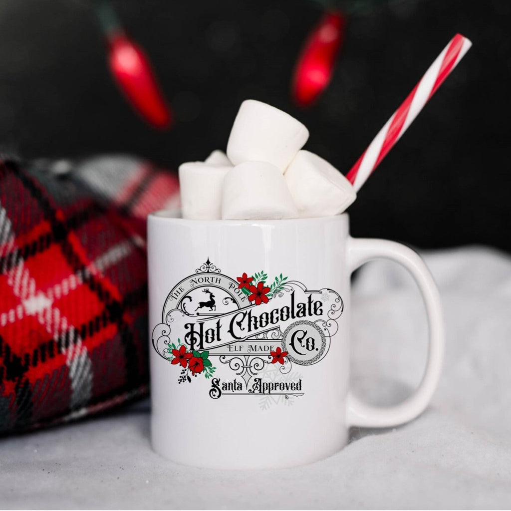 White mug with hot chocolate and marshmallows, featuring a decorative label, against a festive background.