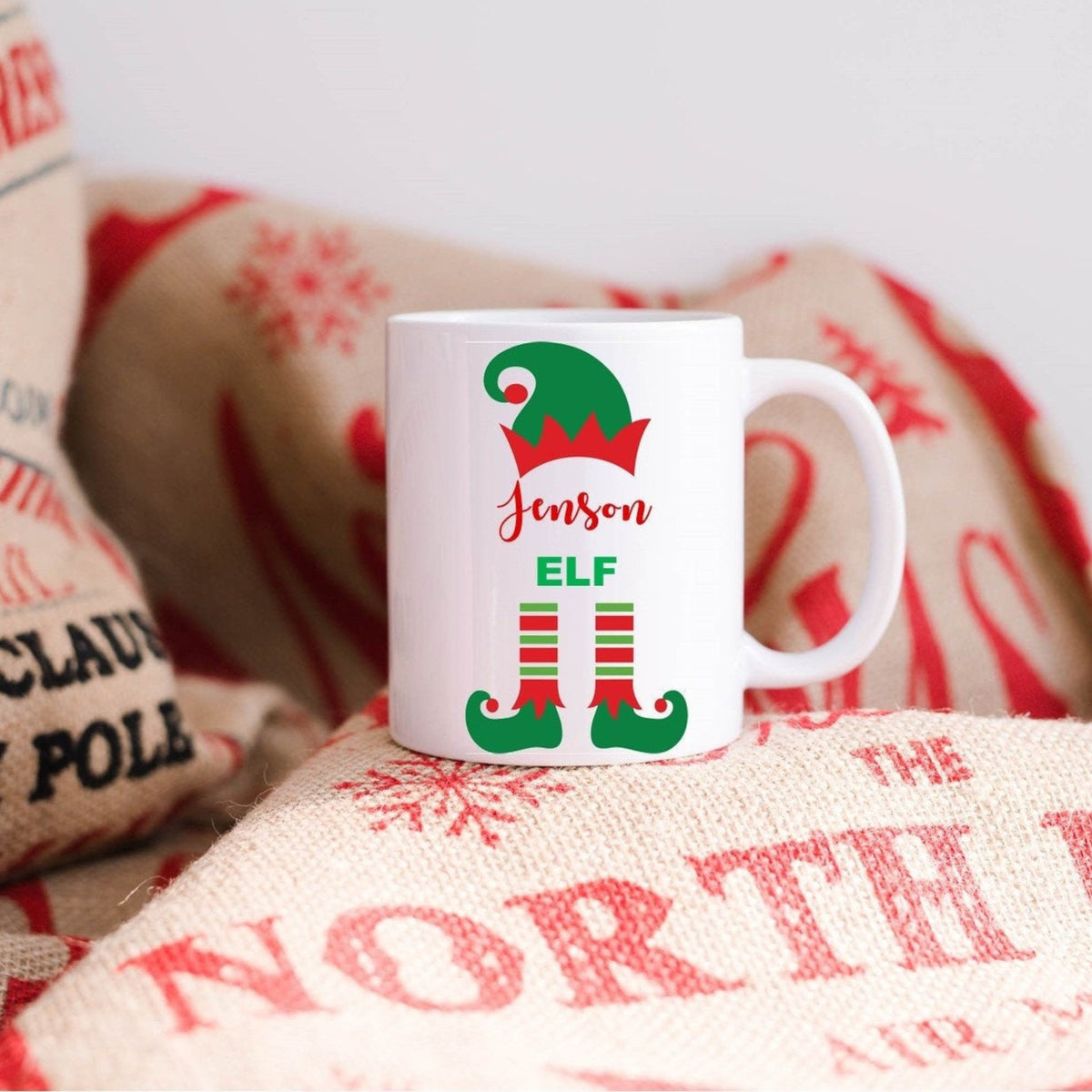 White mug with elf design and text on a textured surface with Christmas-themed sacks.