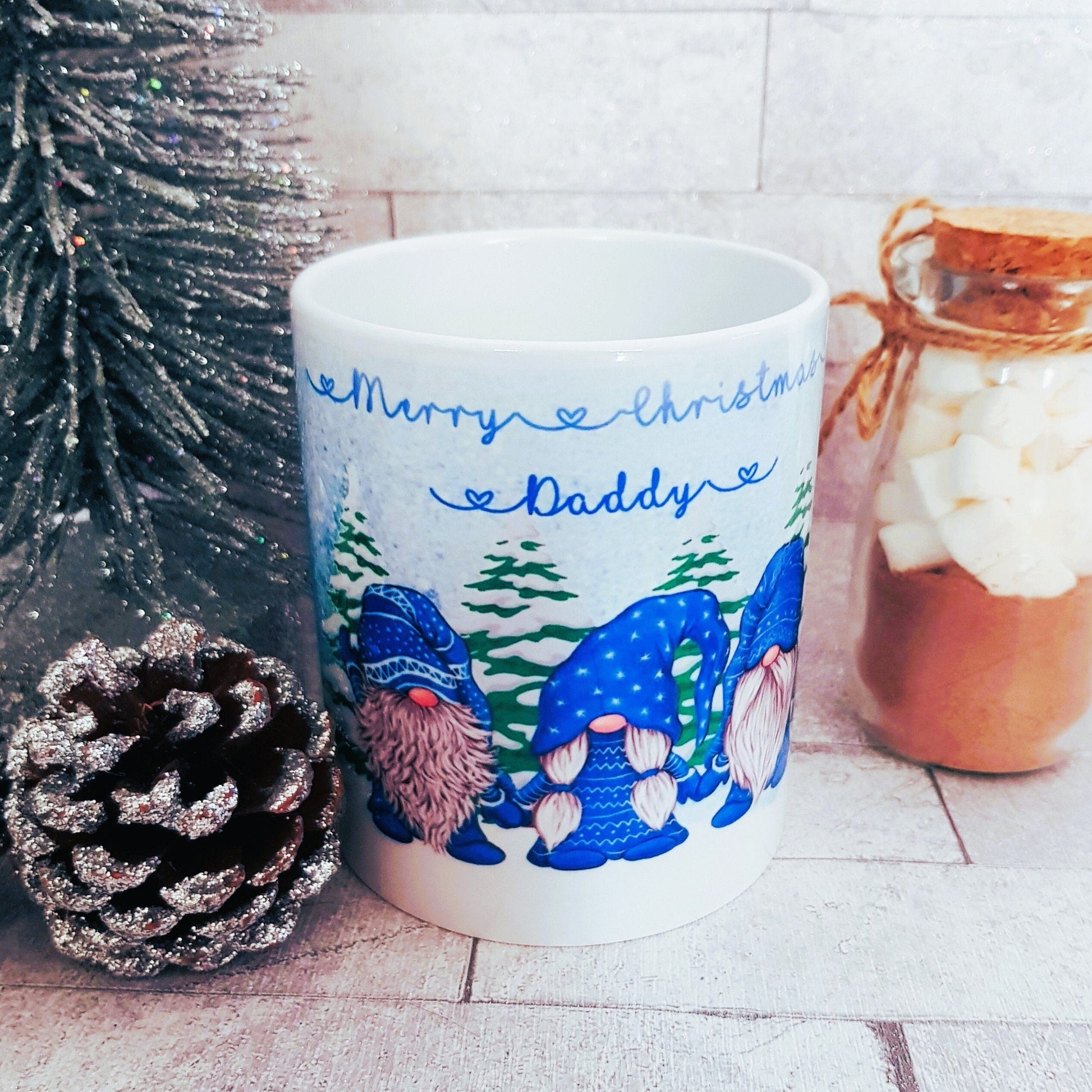 White mug with Christmas-themed design featuring gnomes and text, placed on a surface with decorative items.