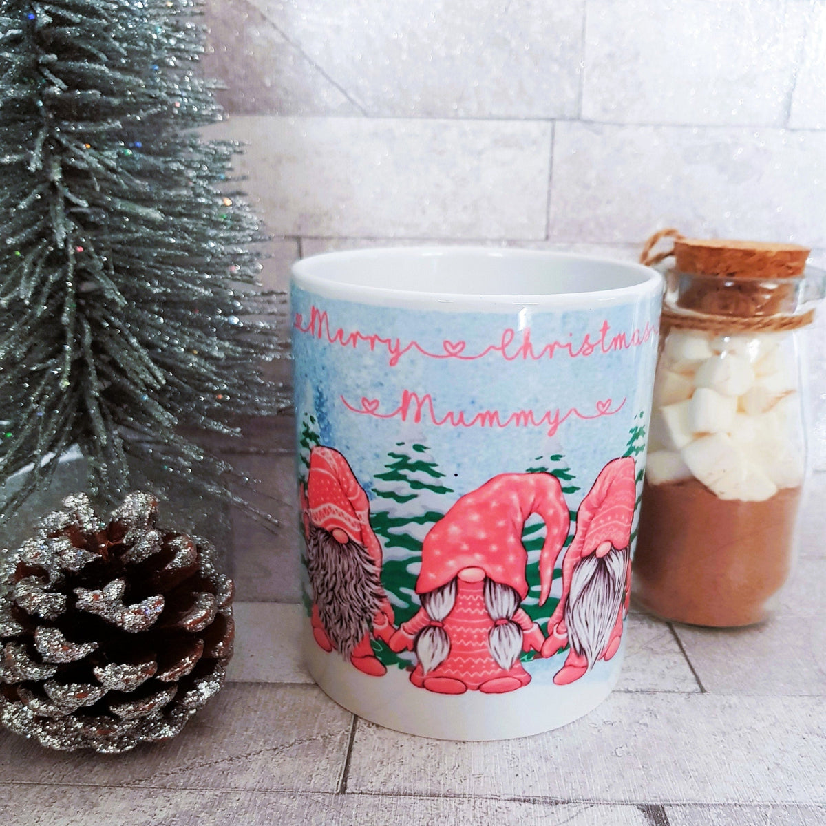 Mug with Christmas design featuring gnomes and text, placed on a surface with decorative elements.