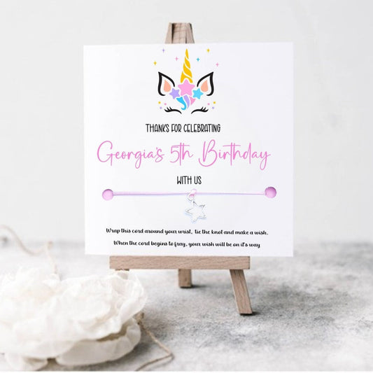 A personalized unicorn-themed wish bracelet displayed on a small wooden easel with 'Thank you for celebrating George's 5th Birthday with us' printed on the card.
