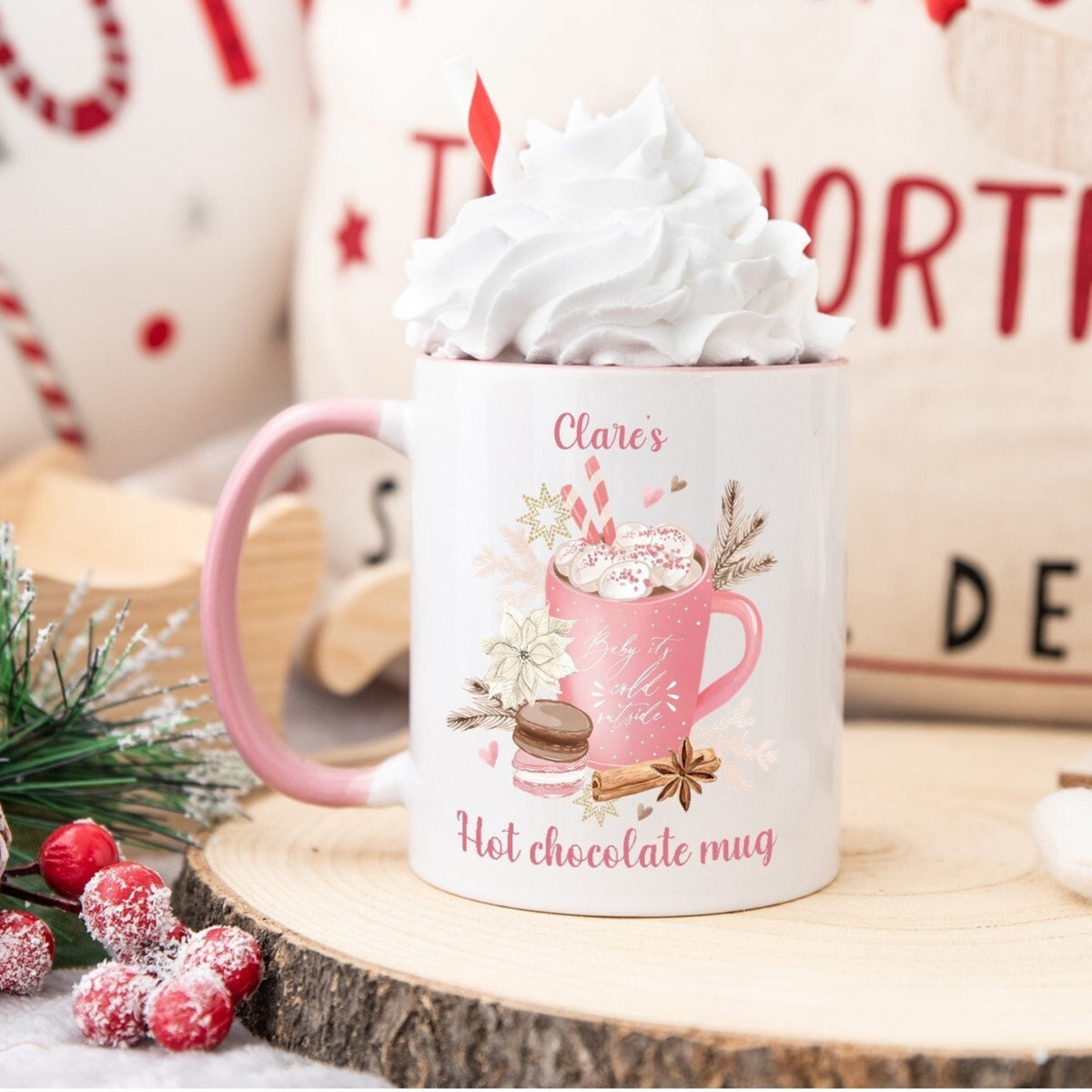 White mug with pink handle and hot chocolate design, topped with whipped cream, on a wooden surface with Christmas decorations.