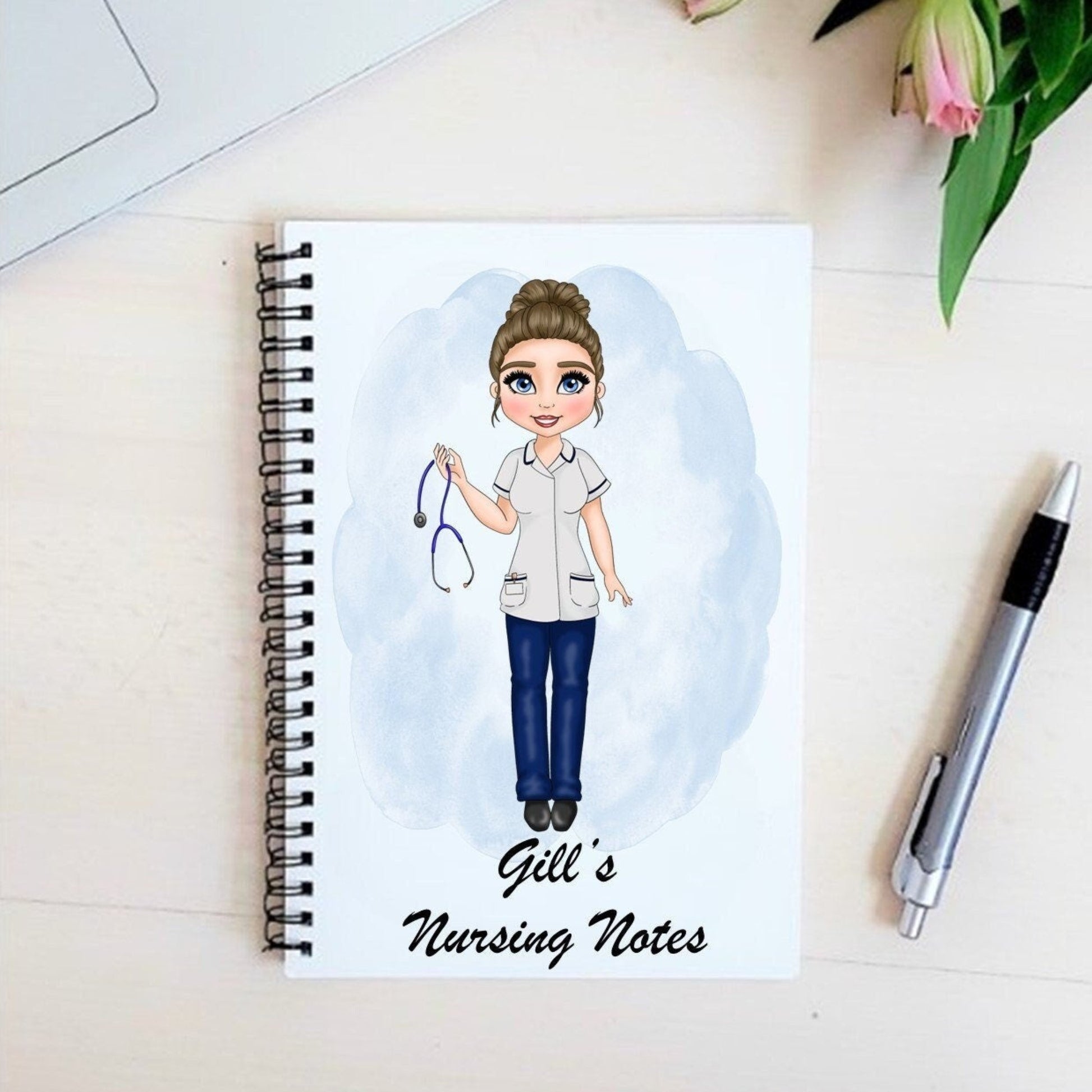 Personalized nursing notebook with illustration of a nurse character on the cover, labeled 'Gill's Nursing Notes', placed on a desk next to a laptop and a pen.
