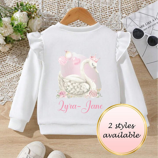 A pink sweatshirt with a swan design and frilled sleeves, personalized with the name 'Lyra Jane', displayed on a wooden hanger next to a pair of white sneakers and a floral arrangement.