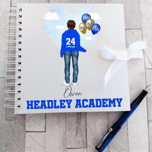 A personalized white scrapbook with a school leaver's autographs, featuring a school girl figure in blue, balloons, and the school name 'Headly Academy' printed on the cover. A blue pen and ribbon are also visible.