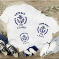 Two white t-shirts with navy anchor designs and text on a wooden surface with shoes and jeans.