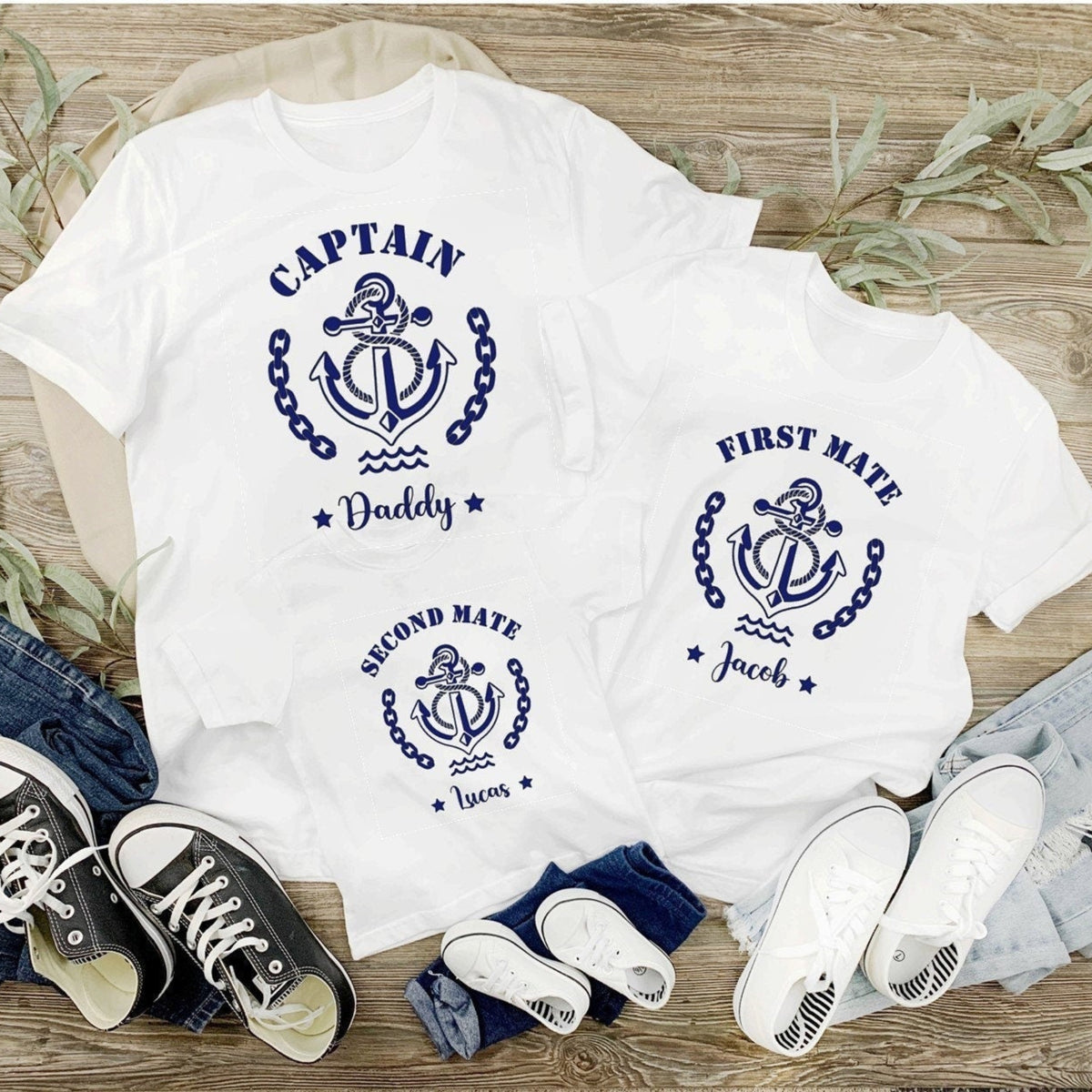 Two white t-shirts with navy anchor designs and text on a wooden surface with shoes and jeans.