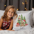Young girl in a checkered dress lying on a bed with a decorative pillow featuring a teddy bear and Christmas trees.