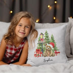 Young girl in a checkered dress lying on a bed with a decorative pillow featuring a teddy bear and Christmas trees.