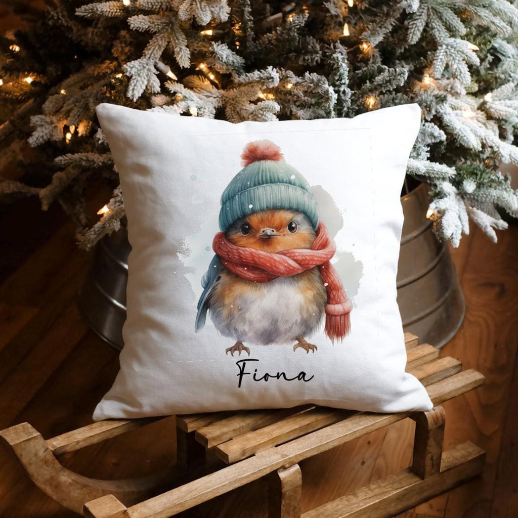 Decorative pillow with a robin design placed on a wooden surface with a Christmas tree in the background. 