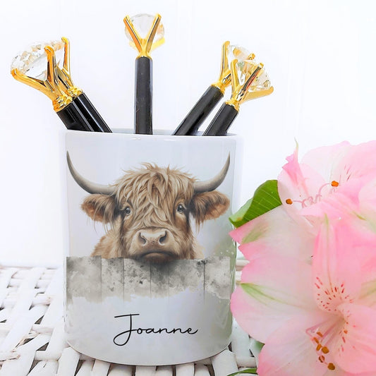Pencil holder with a highland cow design and the name 'Joanne', surrounded by pink flowers on a white surface.