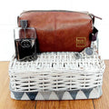 Brown leather toiletry bag with a glass and other items in a white wicker basket on a wooden surface.