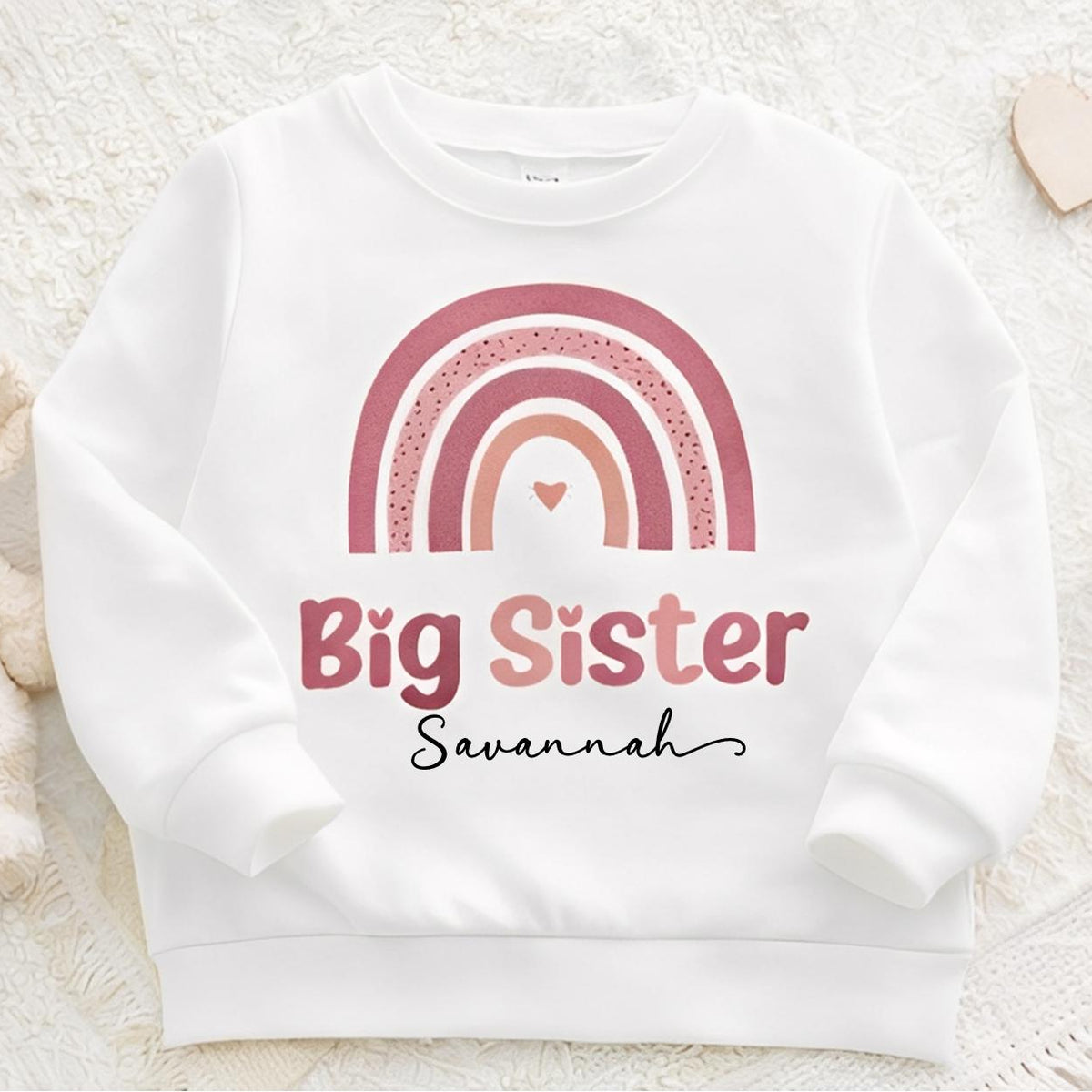 White sweatshirt with pink rainbow and 'Big Sister Savannah' text on a light background with toys.